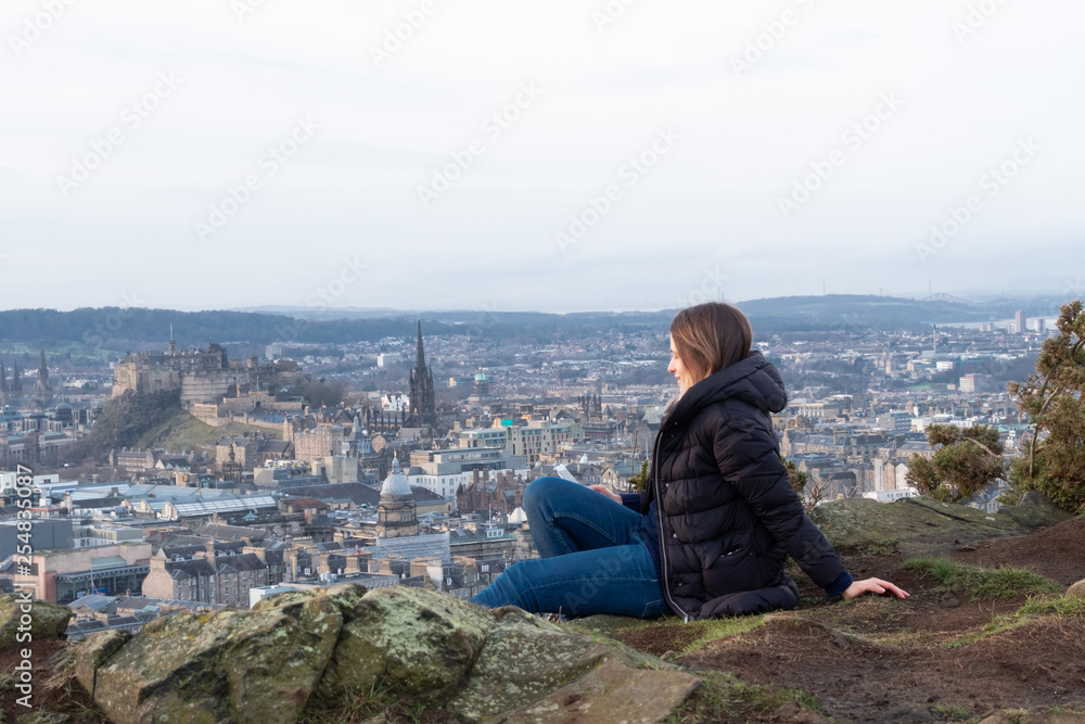 A young woman sits on a high mountain on the background of the city, using the phone