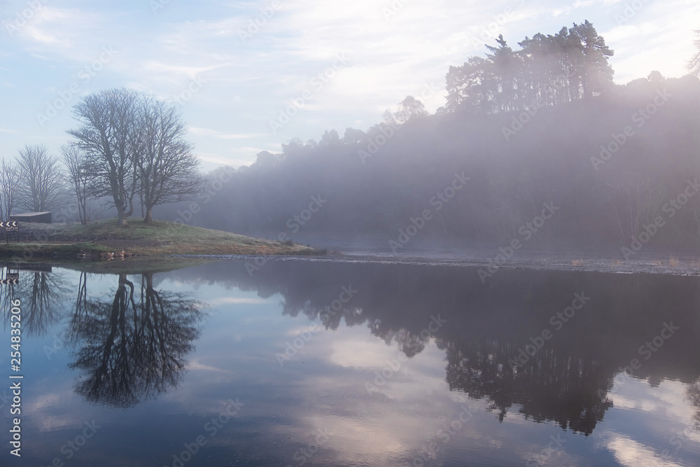 Fototapeta premium A lonely island reflected in the glassy smooth surface of a lake shrouded in morning fog