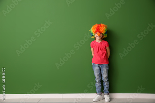 Little girl in rainbow wig and with clown nose near color wall. April fools' day celebration