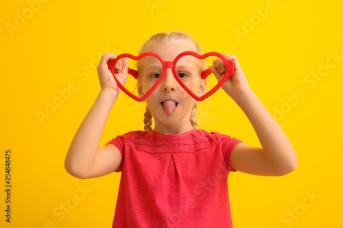 Funny little girl with big heart-shaped glasses on color background. April fools' day celebration