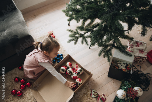 New Year. Decorate the Christmas tree. Artificial tree. A girl in a pink dress with ponytails adorns the Christmas tree. Christmas toys in the box. In anticipation of the holiday.