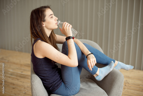Sporty girl with dark hair holds a plastic bottle with clean water in her hands is sitting on a gray chair resting after a workout a healthy diet, a healthy lifestyle
