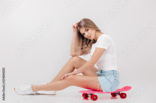 beautiful woman blonde in white t-shirt and denim shorts sitting on pink skateboard. teenager.