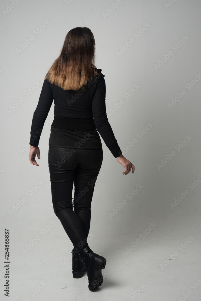 full length portrait of a brunette girl wearing modern black jacket and pants, standing pose with back to the camera on grey studio background. full length portrait of a brunette girl wearing modern black jacket and pants, standing pose with back to the camera on grey studio background.