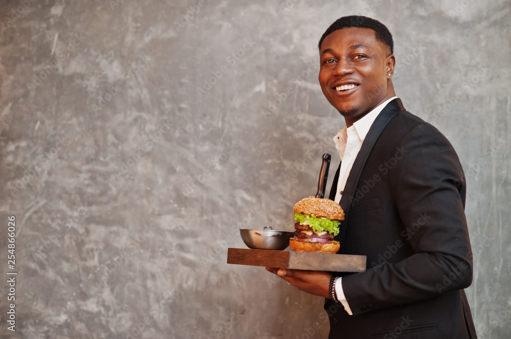 Respectable young african american man in black suit hold tray with double burger against gray wall. Respectable young african american man in black suit hold tray with double burger against gray wall.