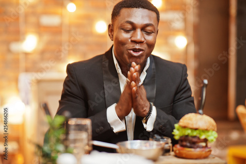 Hungry and happy rubs hands. Respectable young african american man in black suit sitting in restaurant with tasty double burger and soda drink.