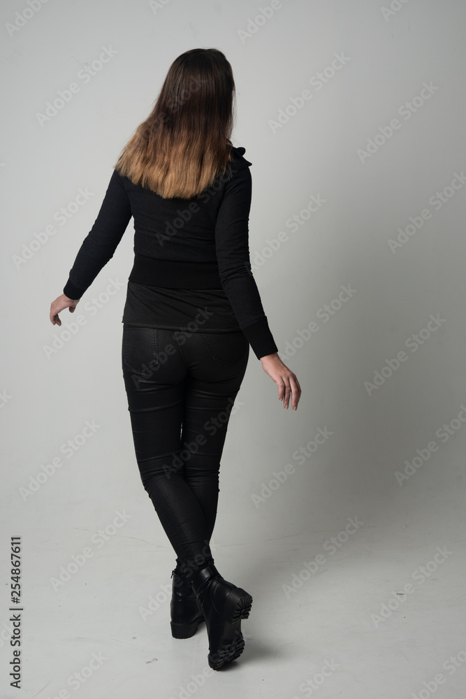 full length portrait of a brunette girl wearing modern black jacket and pants, standing pose with back to the camera on grey studio background. full length portrait of a brunette girl wearing modern black jacket and pants, standing pose with back to the camera on grey studio background.
