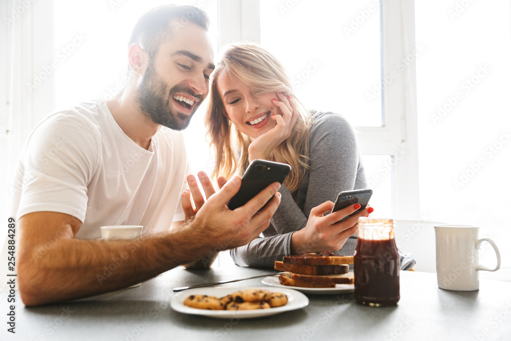 Loving couple sitting at the kitchen have a breakfast using mobile phones. - obrazy, fototapety, plakaty 