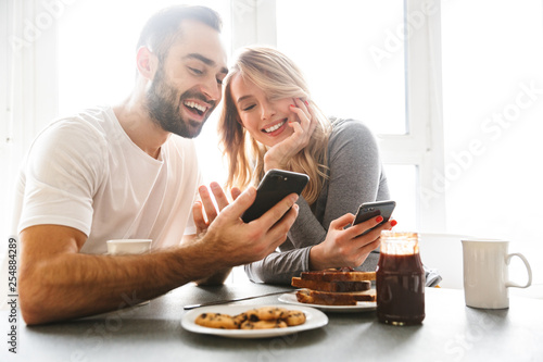 Loving couple sitting at the kitchen have a breakfast using mobile phones.