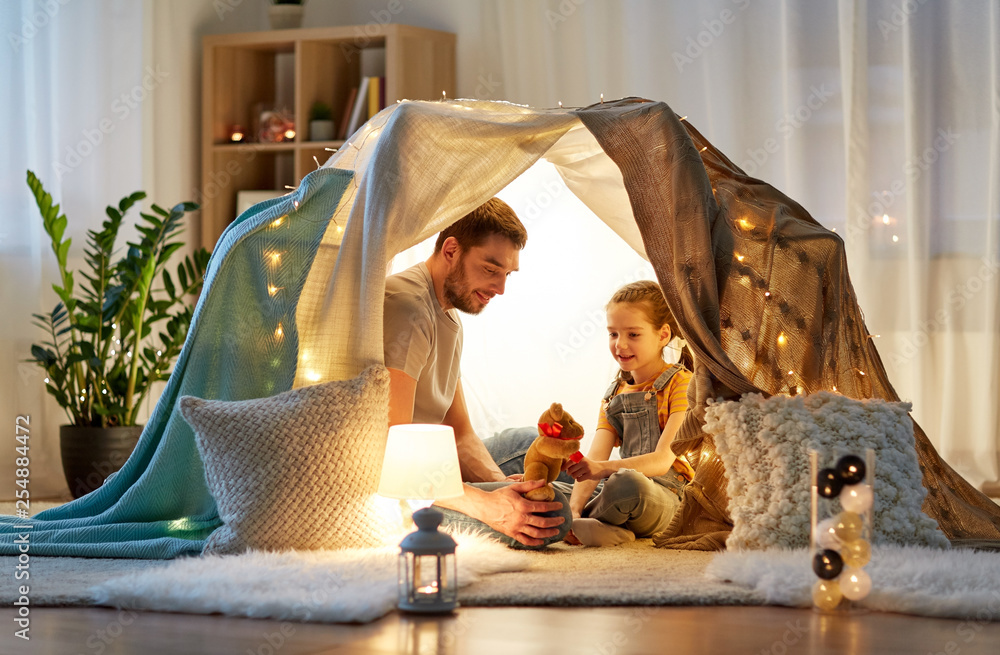 family, hygge and people concept - happy father with teddy bear toy and little daughter playing in kids tent at night at home - obrazy, fototapety, plakaty 