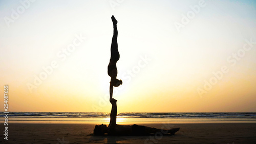 Silhouette of fit sporty couple practicing acrobatic yoga with partner together on the sandy beach. Female acrobat doing handstand on the hands of his lying male partner. 