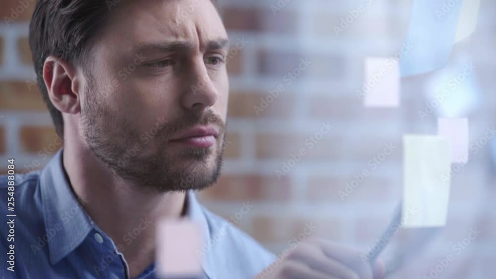 Serious businessman in blue shirt holding pen, reading sticky notes and writing at workplace Serious businessman in blue shirt holding pen, reading sticky notes and writing at workplace
