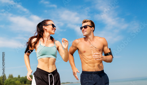 Fototapeta Naklejka Na Ścianę i Meble -  fitness, sport and technology concept - happy couple with earphones and arm bands running along summer beach