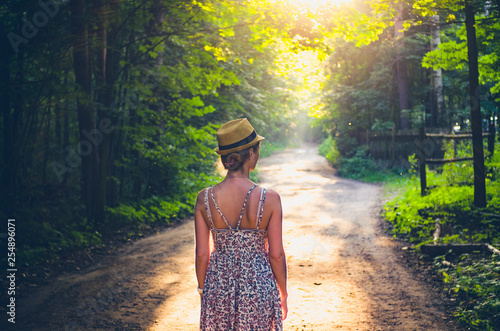 A woman in a straw hat on a street in summer looks thoughtfully at the road ahead. View from the back