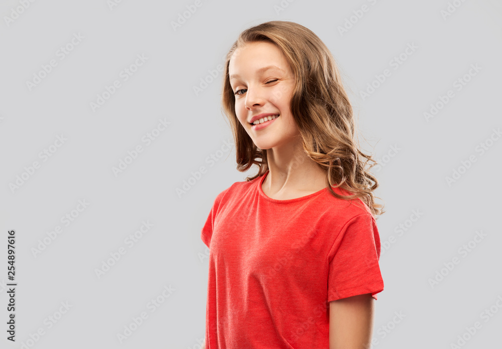 people concept - smiling teenage girl with long hair in red t-shirt winking over grey background people concept - smiling teenage girl with long hair in red t-shirt winking over grey background