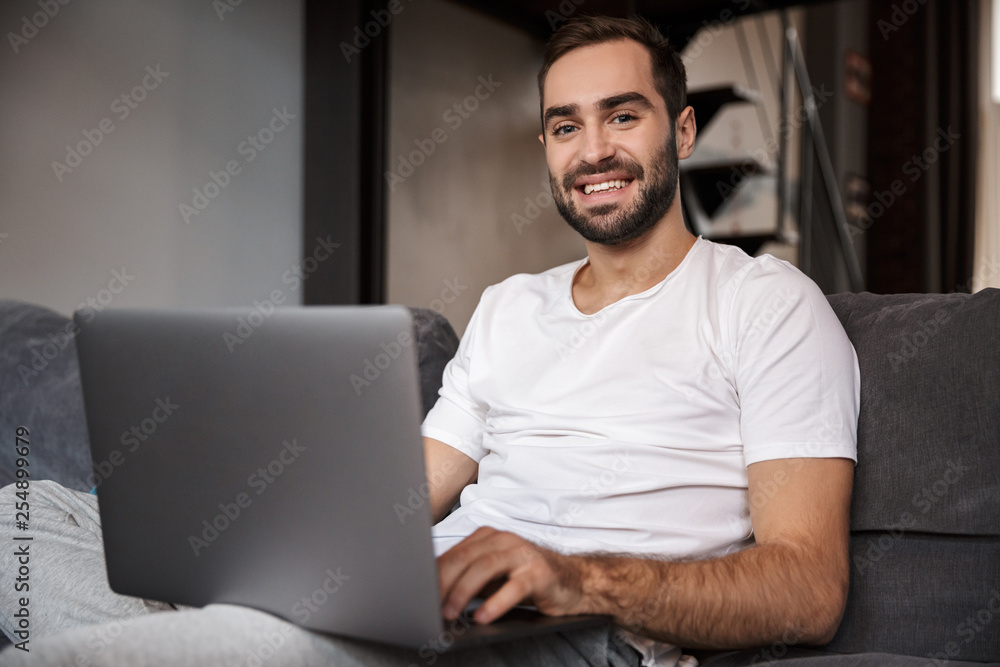 Photo of attractive bachelor using silver laptop while sitting on couch in living room Photo of attractive bachelor using silver laptop while sitting on couch in living room