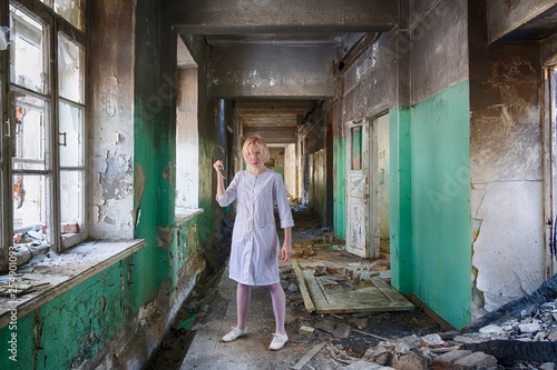 Beautiful woman with knife in white nurse coat (or bathrobe) stand among ruines of abandoned hospital or asylum. Psycho or maniac danger woman