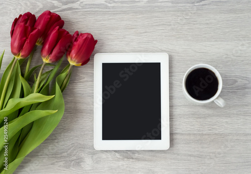 White tablet with a cup of coffee and red flowers lies on a white wooden table.