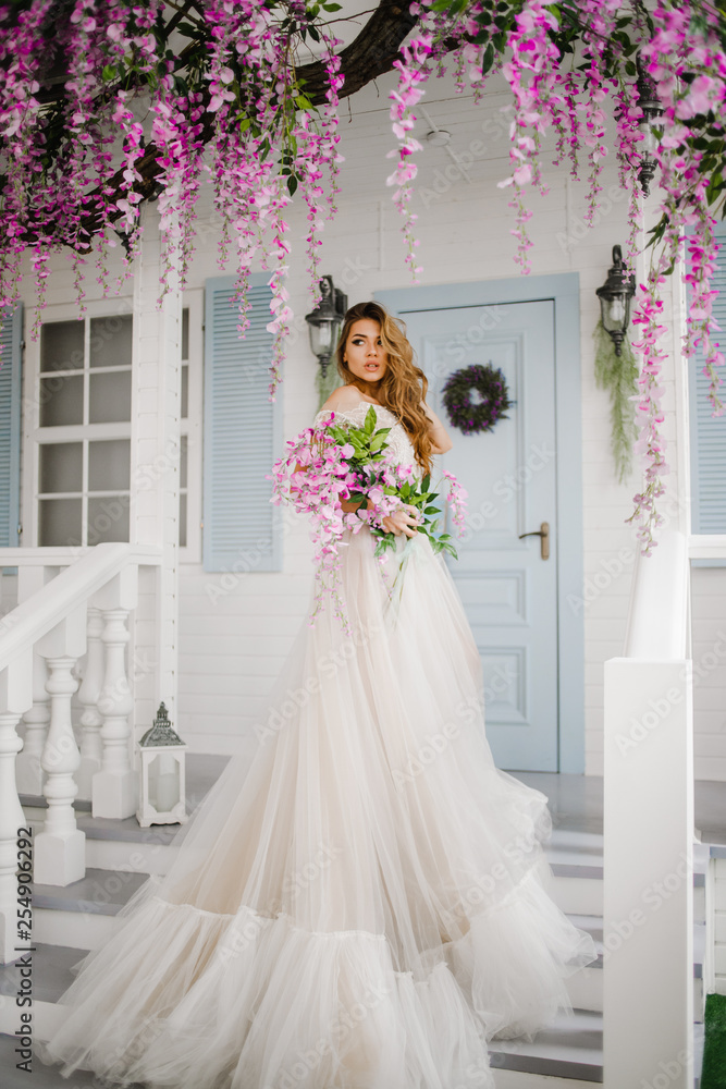 Fototapeta premium Luxury girl in a long white dress standing on the stairs with a bouquet in her hands