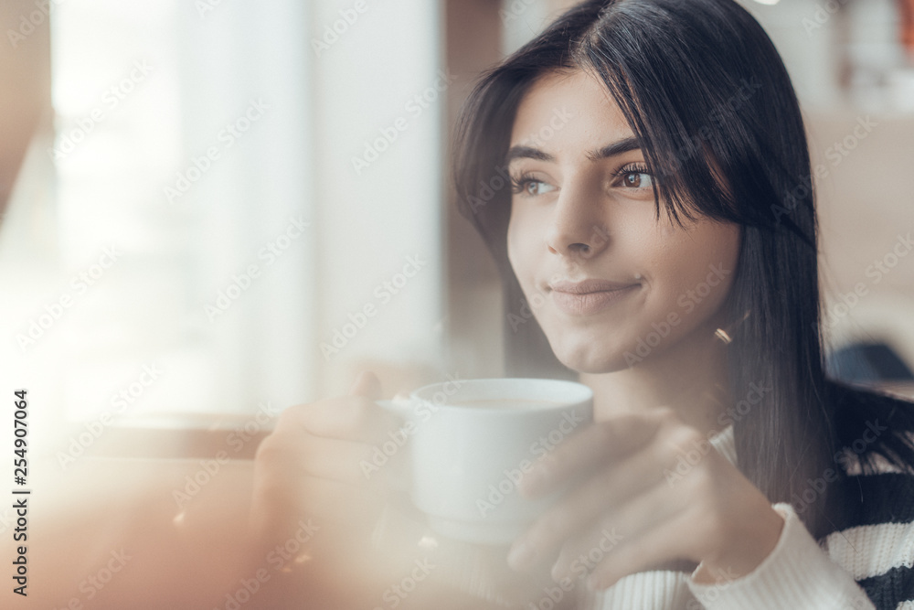 Beautiful caucasian girl in casual clothes with coffe in hands smiling while sitting in cafe. success. Close up, with light leaks Beautiful caucasian girl in casual clothes with coffe in hands smiling while sitting in cafe. success. Close up, with light leaks