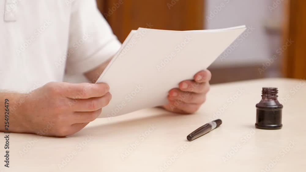 Man piles together white blanks with written letters, puts papers down on beige desk and goes away. Close shot. Side view. Inkpot and brush pen on desk. Selective soft focus. Blurred background Man piles together white blanks with written letters, puts papers down on beige desk and goes away. Close shot. Side view. Inkpot and brush pen on desk. Selective soft focus. Blurred background