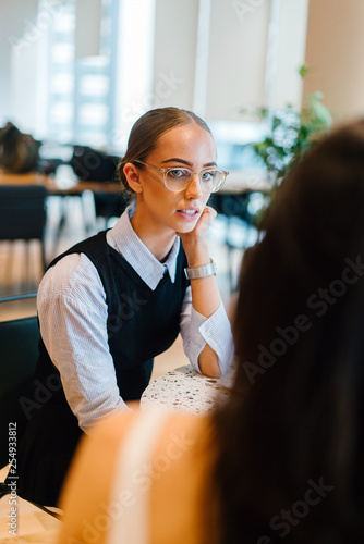 A beautiful Caucasian lady looking at her teammate inside a cafe. She is wearing a nice corporate dress with an expensive watch and eyeglasses.