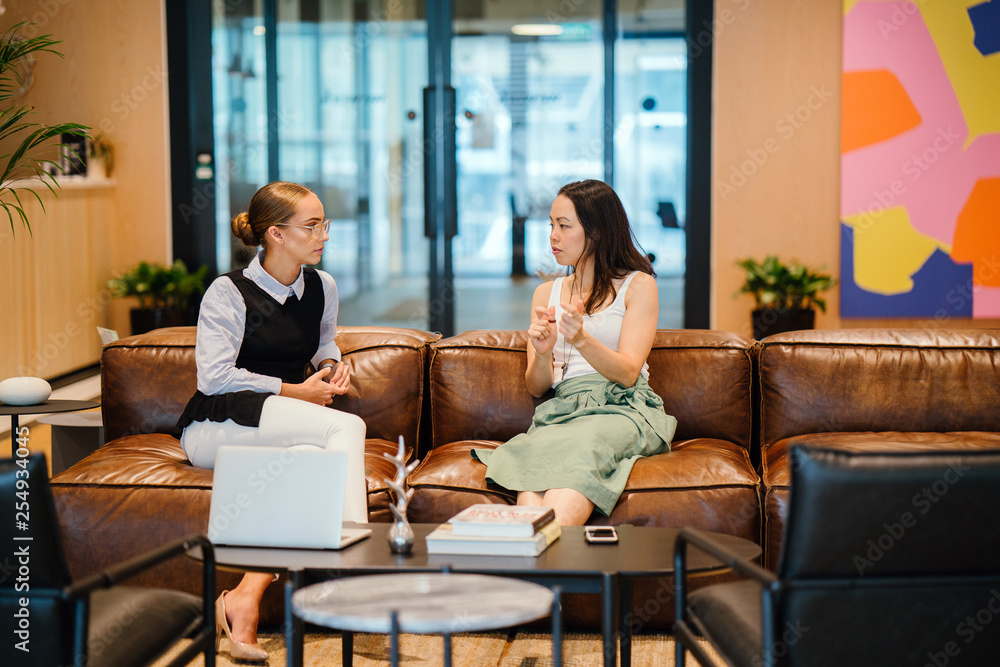 Two delightful women talking about a record inside a bistro. They are discussing some activities while sitting on a brown sofa.