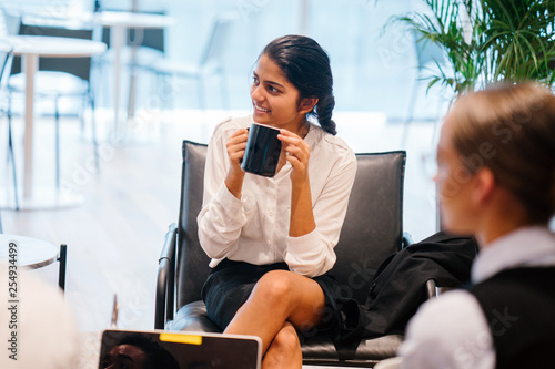A beautiful Indian woman is chatting with a colleague inside a cafe. She is listening to a discussion while drinking hot coffee.