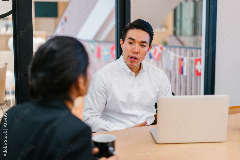 Obraz premium A handsome Asian boss discussing some material with a teammate inside the office. He is looking good in his white polo.