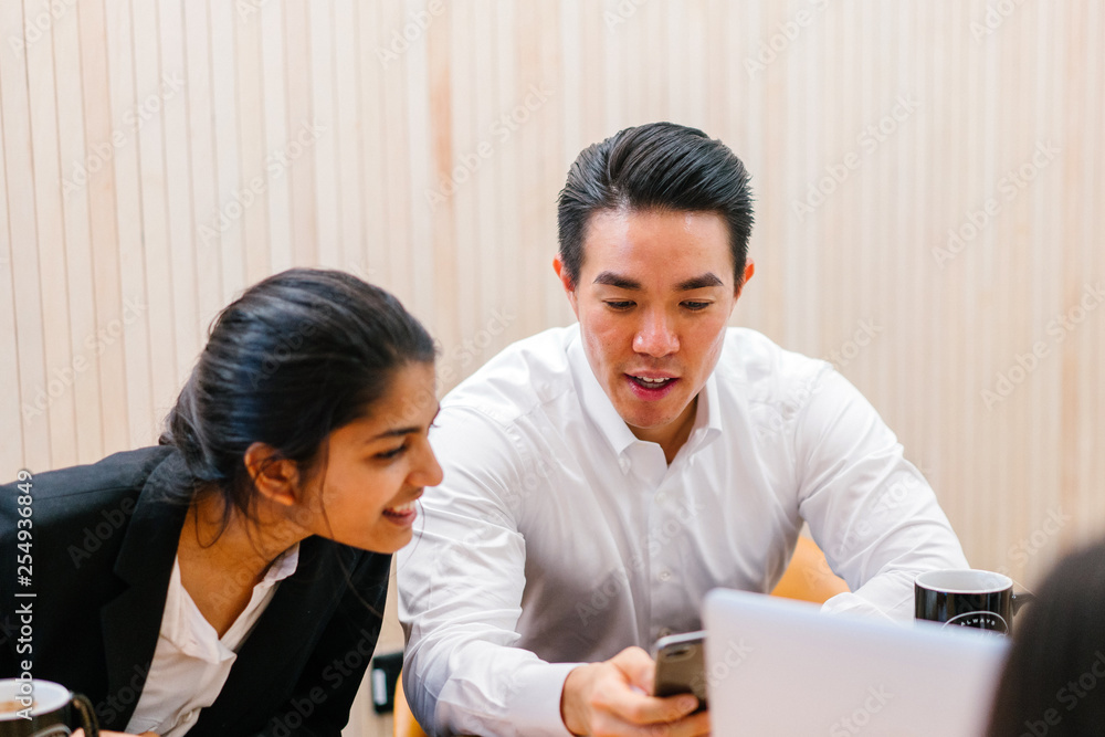 A Chinese Asian manager is showing his cellular phone with his officemate in a meeting room. He is smiling confidently as he talk to his Indian woman colleague who is sitting beside him A Chinese Asian manager is showing his cellular phone with his officemate in a meeting room. He is smiling confidently as he talk to his Indian woman colleague who is sitting beside him