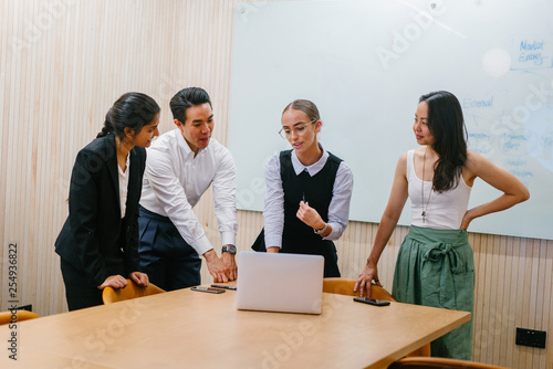 A diverse team stand around a laptop for a discussion in a meeting room in the office during the day. They are ethnically diverse and includes a Caucasian, Indian and Eurasian woman and a Chinese man. photo