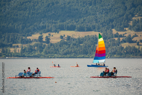 olorful sailboat with rainbow color stripes, a couple of kayaks and family boats. Groups of locals or tourists enjoying. Sailing in the cold waters of the Villarrica lake in a warm summer sunny day