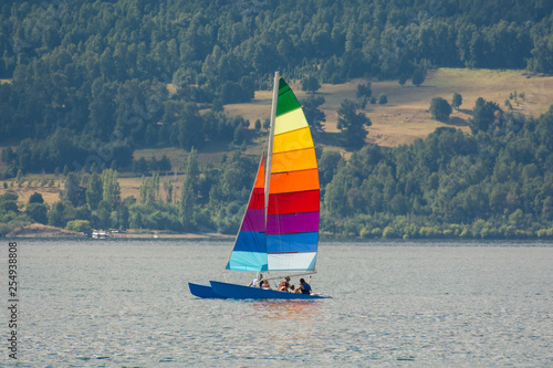 Colorful sailboat with rainbow color stripes, with a group of locals or tourists enjoying, with mountains and forests on the background. Sailing in Villarrica lake in a warm summer sunny day