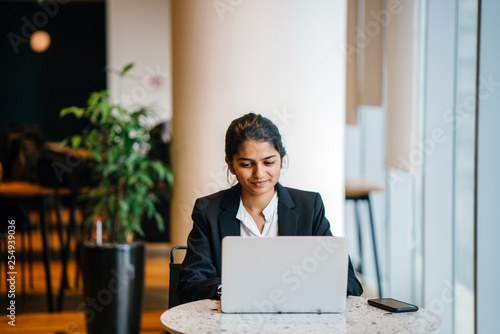 A wonderful Indian lady sitting inside a cafeteria while working. She is smiling in front of a laptop while discussing with a teammate. 