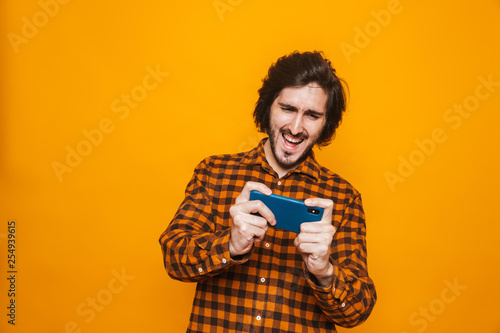 Image of smiling man wearing plaid shirt playing video games on smartphone while standing isolated