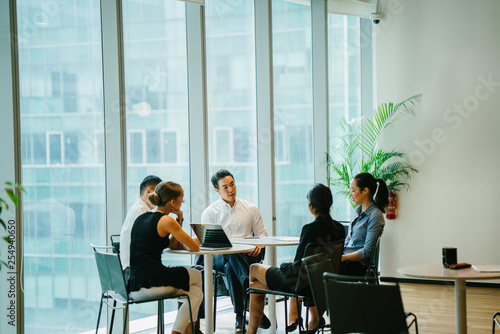 An assorted group lounge around a table in a gathering space to have a conference to talk about plans. The gathering is global with Asian and white colleagues and they are expertly dressed.