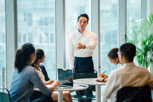 A gorgeous, sure, and fit Asian Chinese man seats a gathering with his group amid the day in the workplace. He is expertly wearing a shirt and pants and is motioning as he talks.