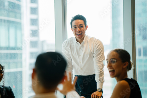 An Asian Chinese businessman is presenting to his colleagues in a conference room. He is wearing a white shirt and black pants.