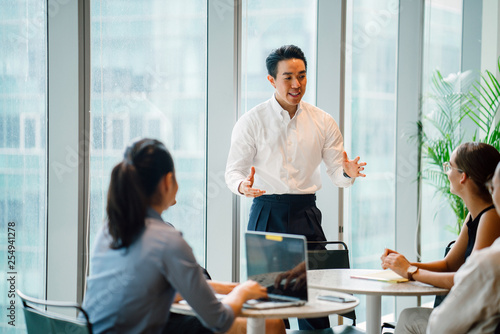 A good looking and confident Asian Chinese man is presenting in a meeting with his team during the day in the office. He is professionally dressed in a polo and pants and is gesturing as he speaks.