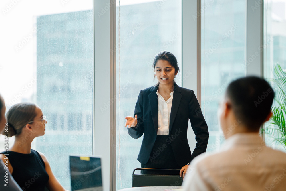 An Asian Indian businesswoman is having a conversation with her colleagues in a conference room while explaining confidently. She is wearing a black blazer and white blouse with a black skirt. An Asian Indian businesswoman is having a conversation with her colleagues in a conference room while explaining confidently. She is wearing a black blazer and white blouse with a black skirt.