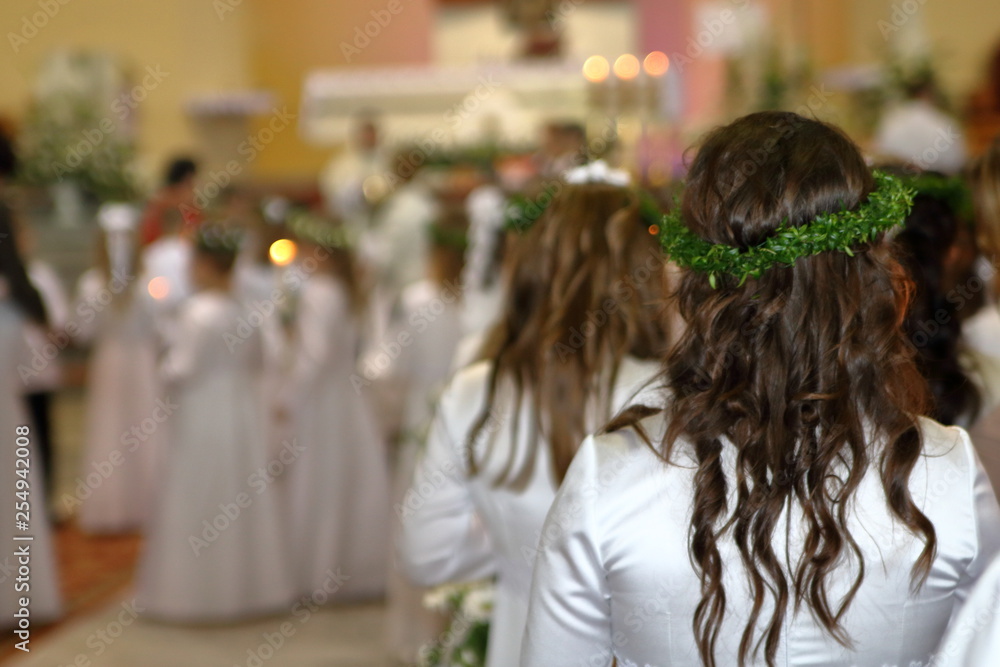 Girls in white occasional dresses in church during First Holy Communion celebration, some kids in soft focus, in background altar, other children in white costumes, priests in soft focus