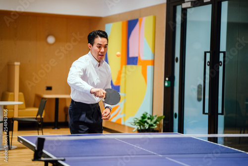 Portrait of a professionally dressed Chinese Asian man playing table tennis with a colleague while having a break in the office during  the day. He is wearing polo with black pants. © Danon