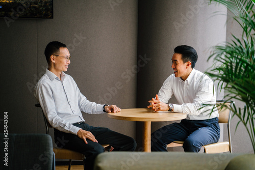 Portrait of two Chinese entrepreneurs sitting at a table. They are smiling as they smile and talk about office matters. Both are wearing white shirt and black pants for the day.