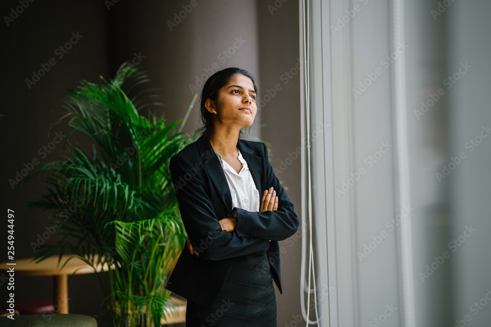 Portrait of a lovely young Indian woman looking at the view from a glass window in her office. She's dressed in a black suit and pants for the day. Portrait of a lovely young Indian woman looking at the view from a glass window in her office. She's dressed in a black suit and pants for the day.