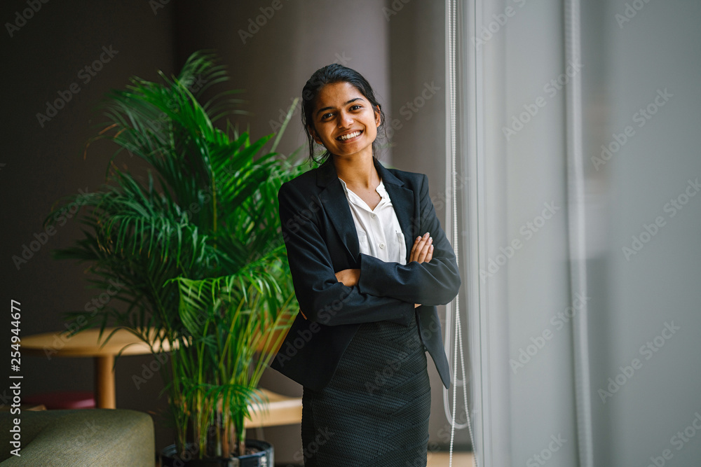 Image of a modern Asian Indian businesswoman standing by a glass window ledge in the office. She laughs and strikes a glamorous pose before the camera.