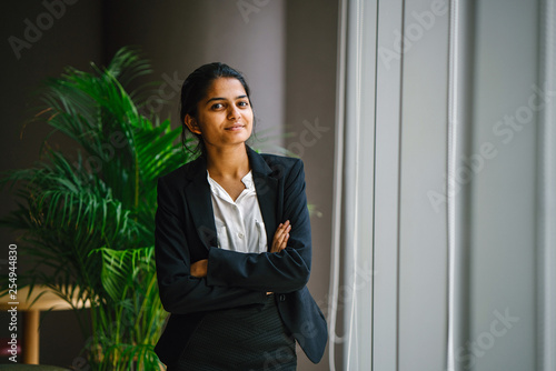 Portrait of a young Asian Indian businesswoman in a meeting room standing by the window, smiling with her arms crossed. She looks optimistic, happy and confident.
