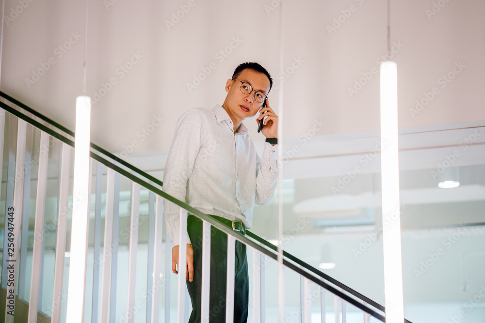 An image of a relaxed and attractive Asian Chinese man walking down the stairs during the day. He is using his smartphone and is wearing a corporate attire. An image of a relaxed and attractive Asian Chinese man walking down the stairs during the day. He is using his smartphone and is wearing a corporate attire.