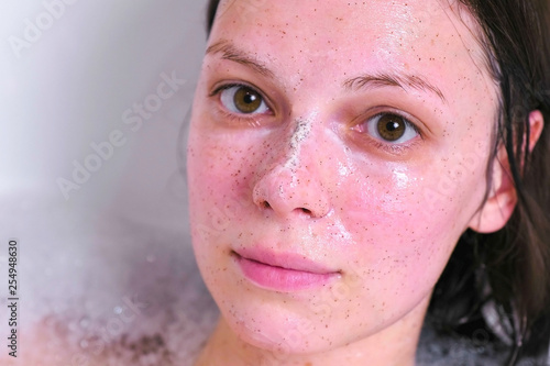 Beautiful brunette woman with scrub on the face lying in the bathroom at home and smiling. Face close-up