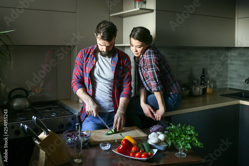 Beautiful young couple in kitchen at home while cooking healthy food. Man is cuts salad. Woman sits on table and watches. Scene from family life.