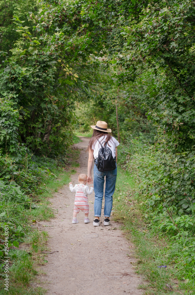 Mom and baby walk walking by the hand on a path in a green park, they go in one direction. View from behind Mom and baby walk walking by the hand on a path in a green park, they go in one direction. View from behind